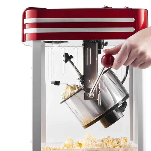 A red retro-style popcorn machine with a clear glass front, shown with a hand operating the metal hopper and lever to pour popcorn into a container below.