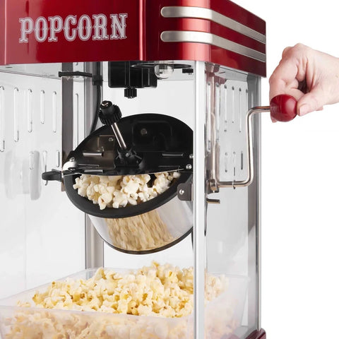A red and silver retro-style popcorn machine with a hand turning the handle, with popcorn popping inside and collected in a clear container below.