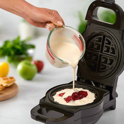 Person pouring batter into a waffle maker with fruit toppings on a kitchen counter.