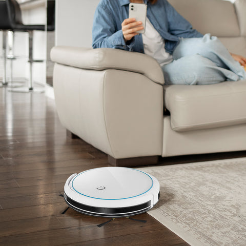 Intellivac 3-in-1 Robot Vacuum cleaner on a wooden floor with a person using a phone on a couch in the background